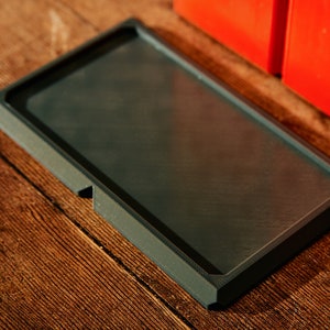 May include: A black rectangular tray with a smooth, slightly textured surface. The tray has a raised edge around the perimeter.