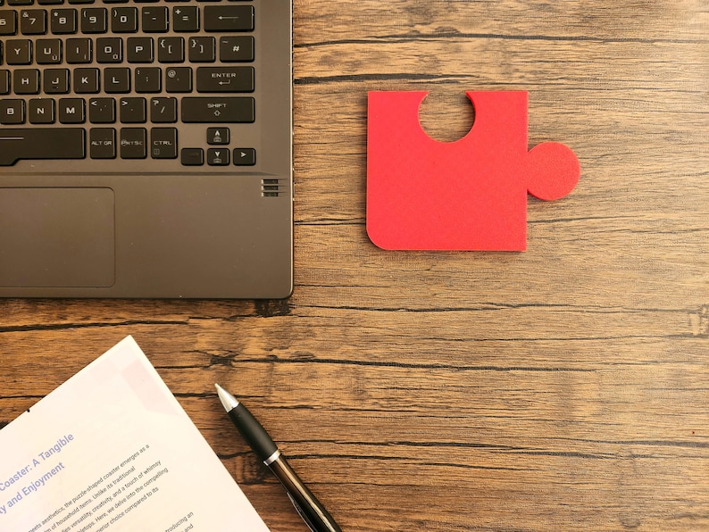 May include: A red puzzle piece on a wooden surface. A laptop, a pen, and a piece of paper with the text "Coaster: A Tangible" are also visible.
