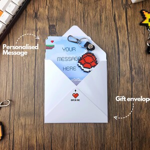 May include: A white envelope with a red heart and the text "Open Me" on the front. The envelope contains a card with a pixelated image of a turtle shell and the text "Your message here". The envelope is accompanied by a keychain with a pixelated image of a gold coin and a pixelated image of a star.