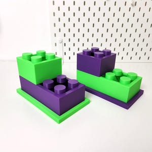 May include: Two sets of stacked toy building blocks. The bottom block in each set is green and the top block is purple. Each block has a series of round studs on top.
