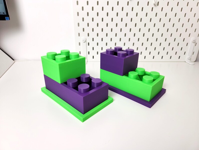 May include: Two sets of stacked toy building blocks. The bottom block in each set is green and the top block is purple. Each block has a series of round studs on top.