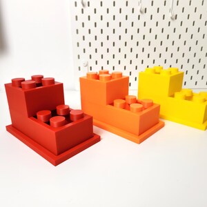 May include: Three 3D printed toy building blocks stacked on top of each other. The bottom block is red, the middle block is orange, and the top block is yellow.