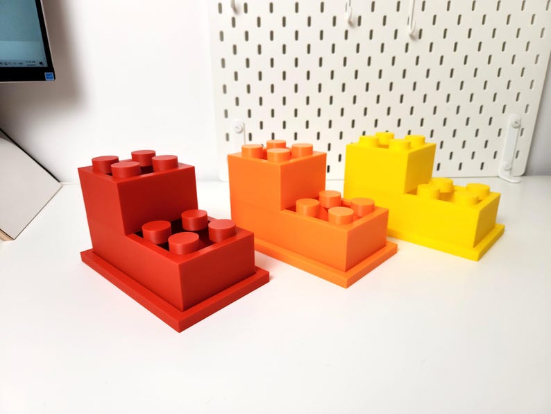 May include: Three 3D printed toy building blocks stacked on top of each other. The bottom block is red, the middle block is orange, and the top block is yellow.