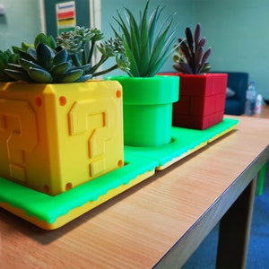 May include: Three potted plants in a green, yellow, and red 3D printed planter set. The planters are shaped like a question mark block, a pipe, and a brick. The plants are succulents.