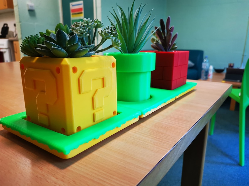 May include: Three potted plants in a green, yellow, and red 3D printed planter set. The planters are shaped like a question mark block, a pipe, and a brick. The plants are succulents.