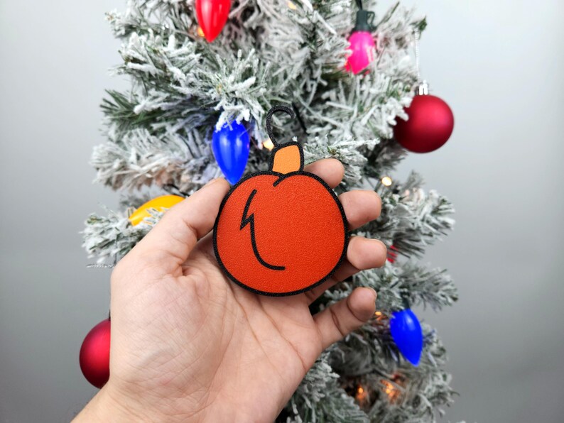 May include: A hand holds a red and orange Christmas ornament shaped like a pumpkin. The ornament has a black outline and a black lightning bolt design.