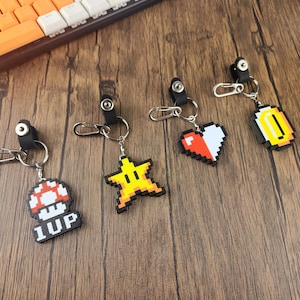 May include: Five keychains with pixelated video game character designs. The keychains feature a red and white heart, a yellow star, a gold coin, a red and white mushroom, and a red and white mushroom with the text "1UP".