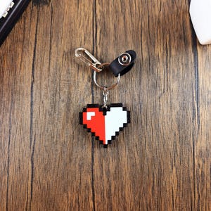 May include: A pixelated heart-shaped keychain with half red and half white. The keychain is attached to a silver ring and a black leather strap.