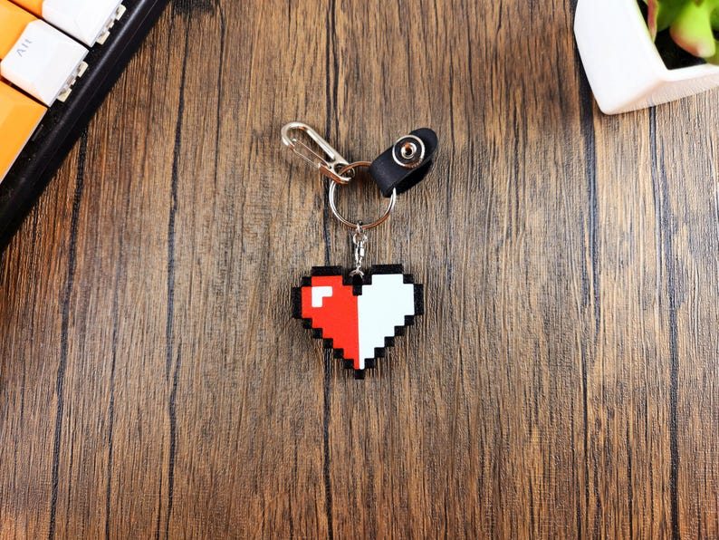 May include: A pixelated heart-shaped keychain with half red and half white. The keychain is attached to a silver ring and a black leather strap.