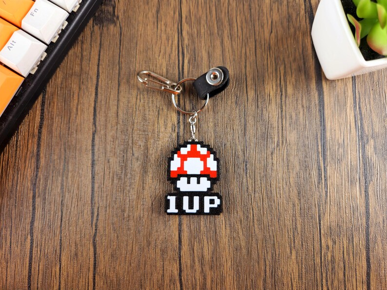 May include: A black and white pixelated mushroom keychain with the word "1UP" on the bottom. The keychain is attached to a silver key ring and a black leather loop.