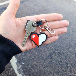 May include: A silver key with a black leather keychain attached to a pixelated heart charm. The heart is half red and half white.