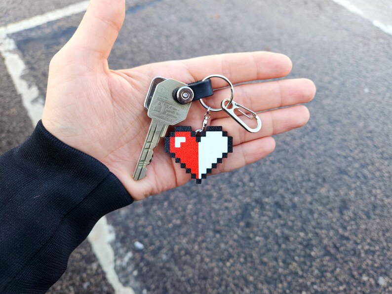 May include: A silver key with a black leather keychain attached to a pixelated heart charm. The heart is half red and half white.