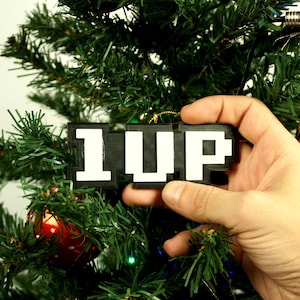 Puede incluir: Un adorno de Navidad pixelado en blanco y negro que dice "1UP" colgado de un &aacute;rbol de Navidad.