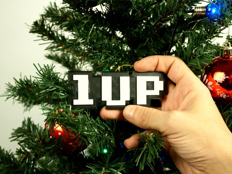 Puede incluir: Un adorno de Navidad pixelado en blanco y negro que dice "1UP" colgado de un &aacute;rbol de Navidad.