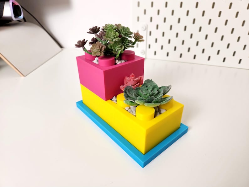May include: Two stacked plastic planters shaped like Lego bricks, the top one is pink and the bottom one is yellow. The planters are filled with small green and red succulents.