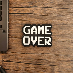 May include: A black and white pixelated sign that reads "GAME OVER" on a wooden surface.