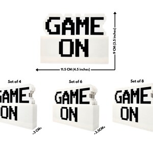 Puede incluir: Un conjunto de letreros blancos pixelados "GAME ON" en tres tama&ntilde;os diferentes. Los letreros est&aacute;n hechos de bloques apilados y tienen 11,5 cm de ancho, 9 cm de alto y 3, 3,5 y 4,5 cm de grosor.