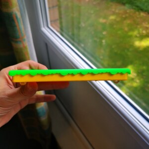 May include: A yellow and green plastic track piece for a toy train set. The track piece has a green grass-like top and a yellow base.