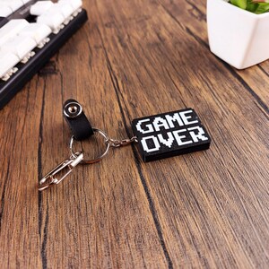 May include: Black keychain with a silver key ring and a black rectangular charm with white pixelated text that reads "GAME OVER".