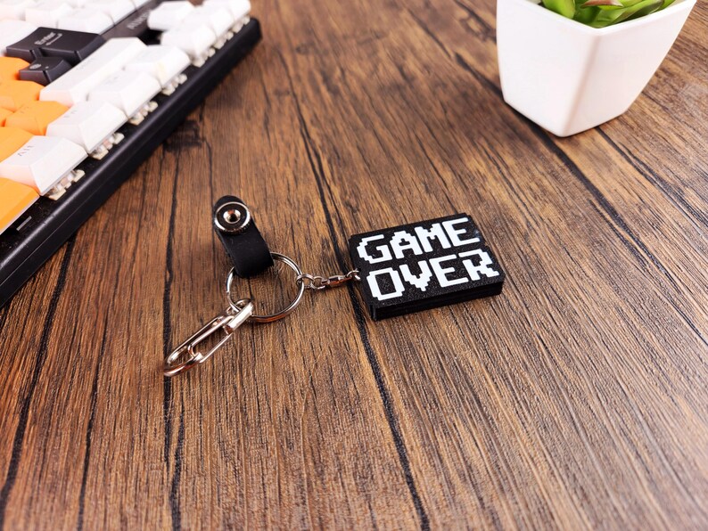 May include: Black keychain with a silver key ring and a black rectangular charm with white pixelated text that reads "GAME OVER".