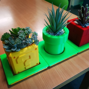 May include: Three potted plants in a green, yellow, and red themed planter set. The planters are shaped like a question mark block, a pipe, and a brick block from the Super Mario Bros. video game.