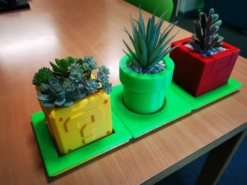 May include: Three potted plants in a green, yellow, and red themed planter set. The planters are shaped like a question mark block, a pipe, and a brick block from the Super Mario Bros. video game.