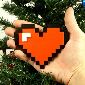 Puede incluir: Un adorno de coraz&oacute;n rojo pixelado con contorno negro, sostenido en una mano contra un fondo de &aacute;rbol de Navidad verde.
