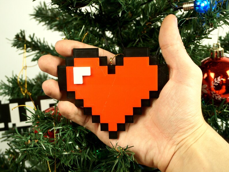 Puede incluir: Un adorno de coraz&oacute;n rojo pixelado con contorno negro, sostenido en una mano contra un fondo de &aacute;rbol de Navidad verde.
