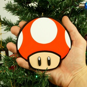 Puede incluir: Un adorno navide&ntilde;o de color rojo y blanco con forma de hongo, con ojos negros y un tallo marr&oacute;n. El adorno se est&aacute; sosteniendo en una mano contra un fondo de &aacute;rbol de Navidad verde.