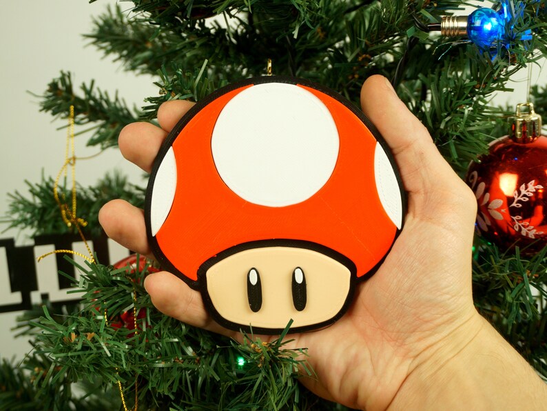 Puede incluir: Un adorno navide&ntilde;o de color rojo y blanco con forma de hongo, con ojos negros y un tallo marr&oacute;n. El adorno se est&aacute; sosteniendo en una mano contra un fondo de &aacute;rbol de Navidad verde.