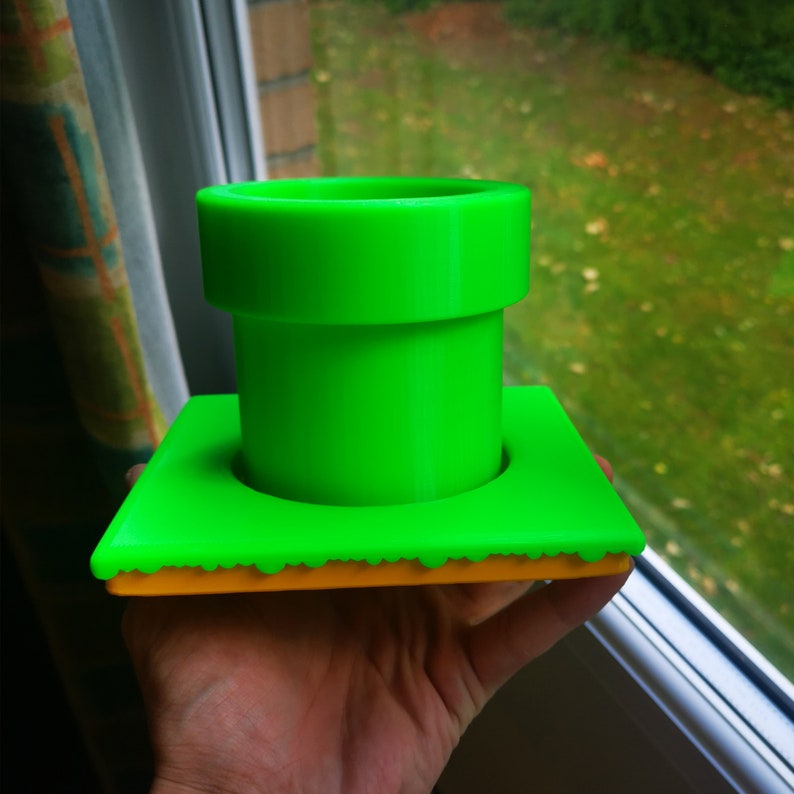 May include: A green plastic pipe-shaped planter with a yellow base. The planter is designed to look like a Super Mario Bros. pipe.