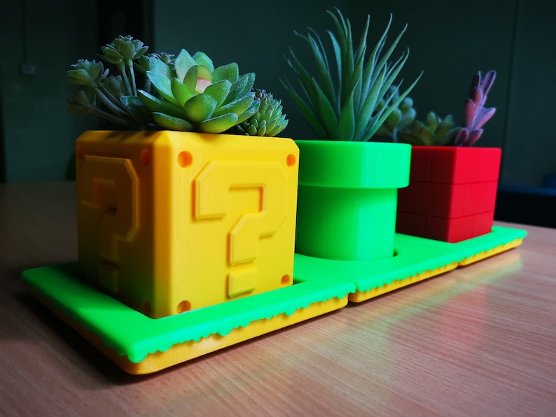 May include: Three 3D printed plant pots shaped like iconic Super Mario Bros. game elements. The pots are yellow with a question mark, green with a pipe, and red with a brick block. Each pot has a small succulent plant growing inside.