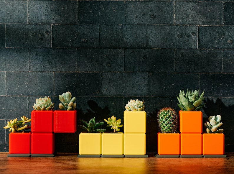 May include: A set of colorful, modular planters shaped like blocks from the classic video game Pac-Man. The planters are red, yellow, and orange and are filled with various succulents and cacti.