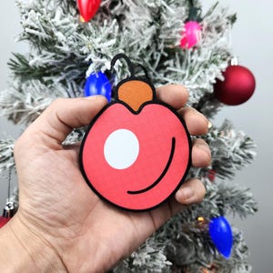 May include: A red and orange Christmas ornament with a white circle in the center. The ornament is being held in a hand.