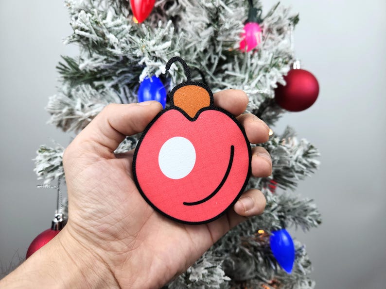 May include: A red and orange Christmas ornament with a white circle in the center. The ornament is being held in a hand.