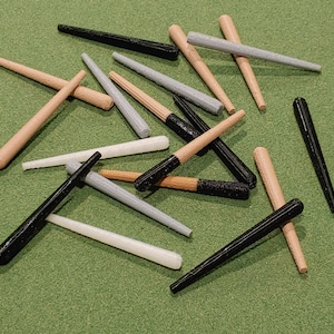 Puede incluir: Una colección de clavijas de madera con detalles de pintura negra, gris y blanca. Las clavijas están dispuestas en un patrón aleatorio sobre una superficie verde.