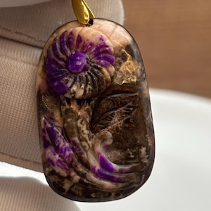 Puede incluir: Un colgante de piedra tallada de color morado y marr&oacute;n con un dise&ntilde;o de flor. El colgante est&aacute; unido a una cadena de oro.