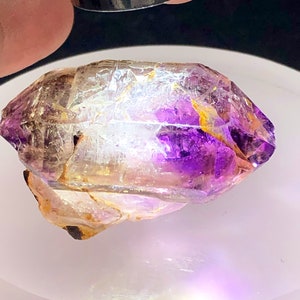 May include: A clear quartz crystal with purple amethyst inclusions. The crystal is faceted and has a pointed tip. It is a natural gemstone.