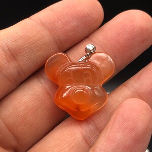 K&ouml;nnte beinhalten: Orangener Anh&auml;nger in Form einer Zeichentrickmaus. Der Anh&auml;nger hat eine silberfarbene &Ouml;se. Das Gesicht der Maus ist mit geschnitzten Merkmalen detailliert. Der Anh&auml;nger wird in einer Hand vor dunklem Hintergrund gehalten.