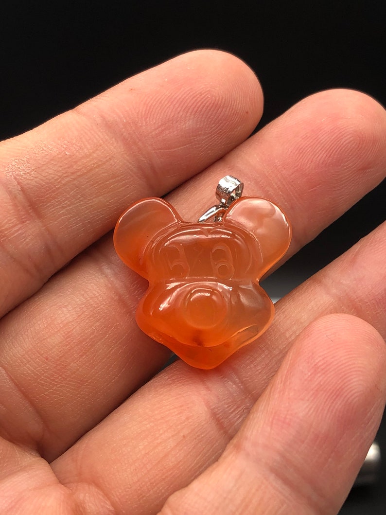 K&ouml;nnte beinhalten: Orangener Anh&auml;nger in Form einer Zeichentrickmaus. Der Anh&auml;nger hat eine silberfarbene &Ouml;se. Das Gesicht der Maus ist mit geschnitzten Merkmalen detailliert. Der Anh&auml;nger wird in einer Hand vor dunklem Hintergrund gehalten.