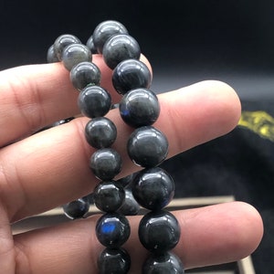 May include: Two beaded bracelets with dark gray, spherical beads. The beads have a polished, reflective surface. The bracelets are held in a hand, showcasing the varying sizes of the beads. The background is dark, emphasizing the bracelets.