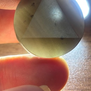 May include: A round, pale green pendant with a triangle design etched into its surface. The pendant has a small hole at the top for a chain. The pendant is held between two fingers.