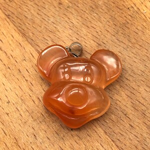 K&ouml;nnte beinhalten: Ein durchscheinender, orangefarbener Anh&auml;nger in Form eines Cartoon-Mauskopfes. Der Anh&auml;nger hat oben eine silberfarbene &Ouml;se zum Anbringen an einer Kette. Die Merkmale der Maus sind deutlich definiert, mit abgerundeten Ohren und einem l&auml;chelnden Gesicht.