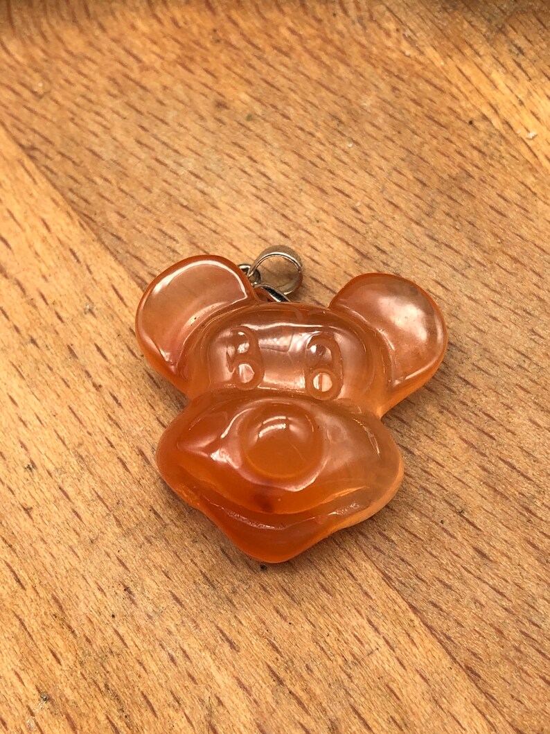 K&ouml;nnte beinhalten: Ein durchscheinender, orangefarbener Anh&auml;nger in Form eines Cartoon-Mauskopfes. Der Anh&auml;nger hat oben eine silberfarbene &Ouml;se zum Anbringen an einer Kette. Die Merkmale der Maus sind deutlich definiert, mit abgerundeten Ohren und einem l&auml;chelnden Gesicht.