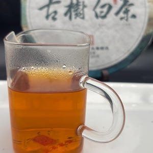 May include: A clear glass mug filled with amber tea, with a handle on the right side. The tea is in front of a round, light-colored tea cake with Chinese characters. The tea has a few small pieces floating in it.