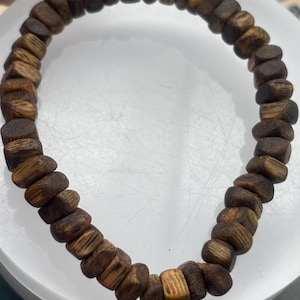 May include: A wooden bead bracelet featuring rectangular and oval-shaped beads in shades of brown and tan. The bracelet is strung together, creating a circular shape. The beads have a natural, textured appearance.