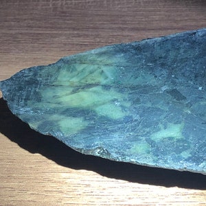 Könnte beinhalten: Ein Stück roher Jadestein mit dunkelgrüner und blauer Farbe. Der Stein hat eine raue, unebene Kante und eine strukturierte Oberfläche. Der Jade wird auf einer Holzoberfläche präsentiert.