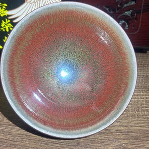 Puede incluir: Un cuenco de té de cerámica con un esmalte degradado que transita desde un centro oscuro y metálico hacia un tono rojo anaranjado vibrante, terminando en un borde gris claro. La superficie interior muestra una textura radial fina.