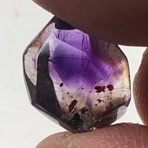 Puede incluir: Un cristal de amatista púrpura con una forma facetada única. El cristal tiene un tono púrpura oscuro con toques de marrón y negro.