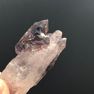 May include: A purple amethyst crystal cluster with a clear quartz point. The amethyst has a deep purple color and is translucent. The quartz point is clear and has a smooth surface.